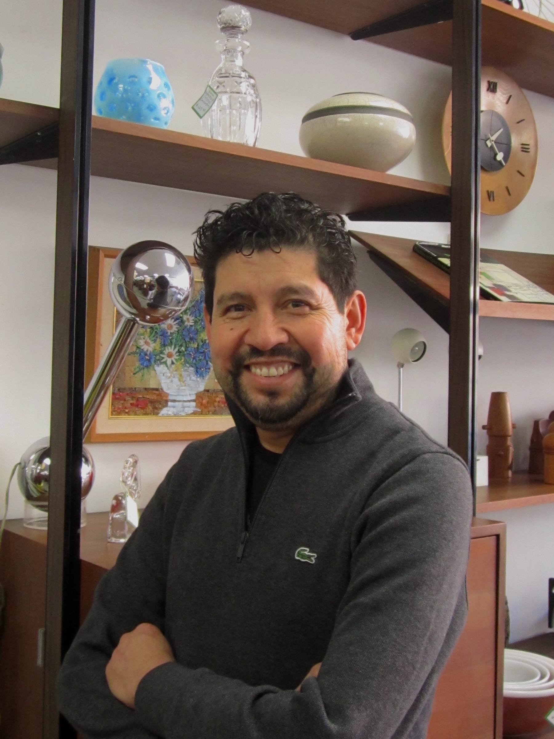 Martin Reyes, owner of Three Stars Vintage, smiling in the shop in front of a shelf of glass and ceramics.