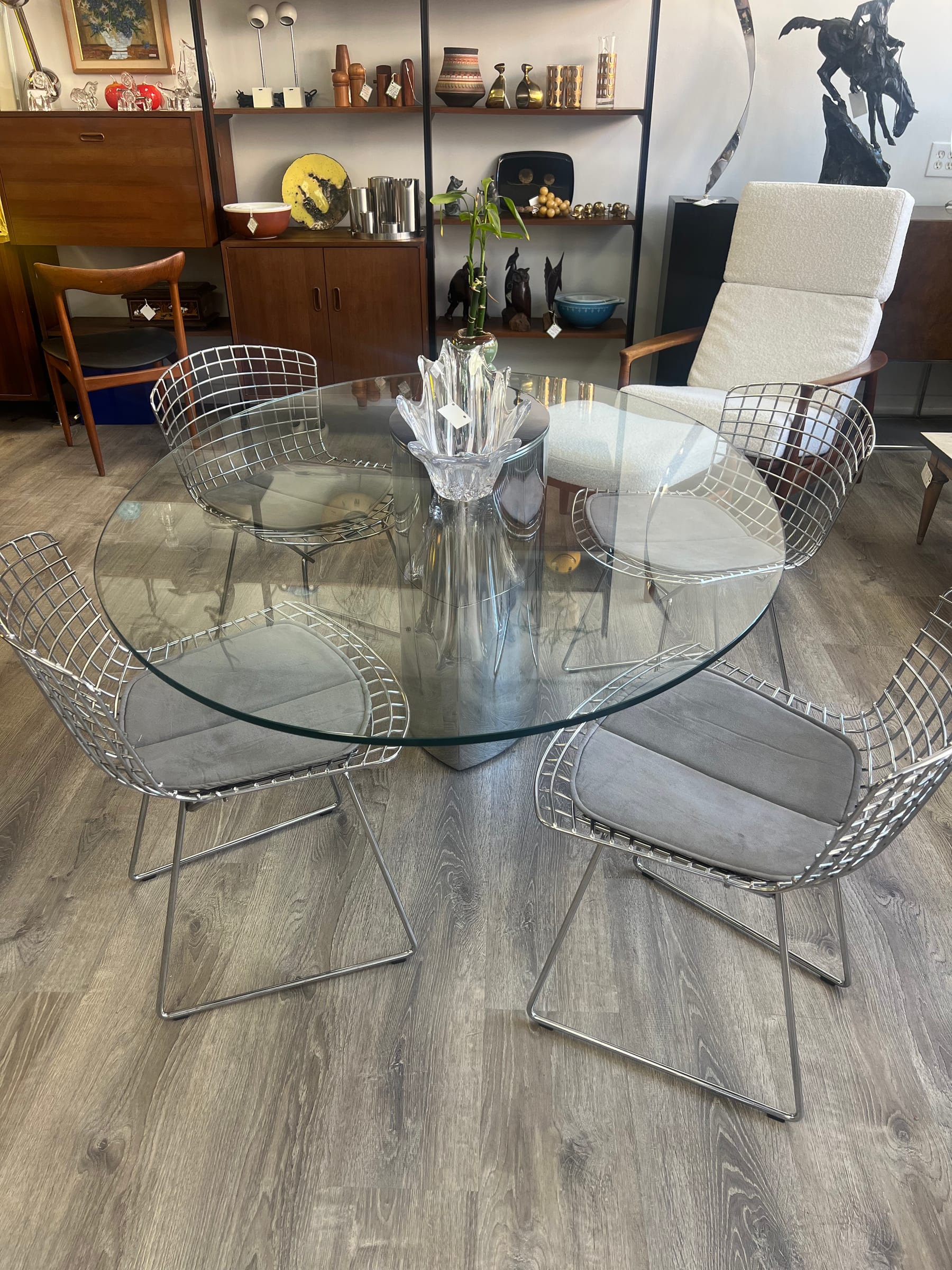 A round glass-top dining table surrounded by four chrome Bertoia side chairs.