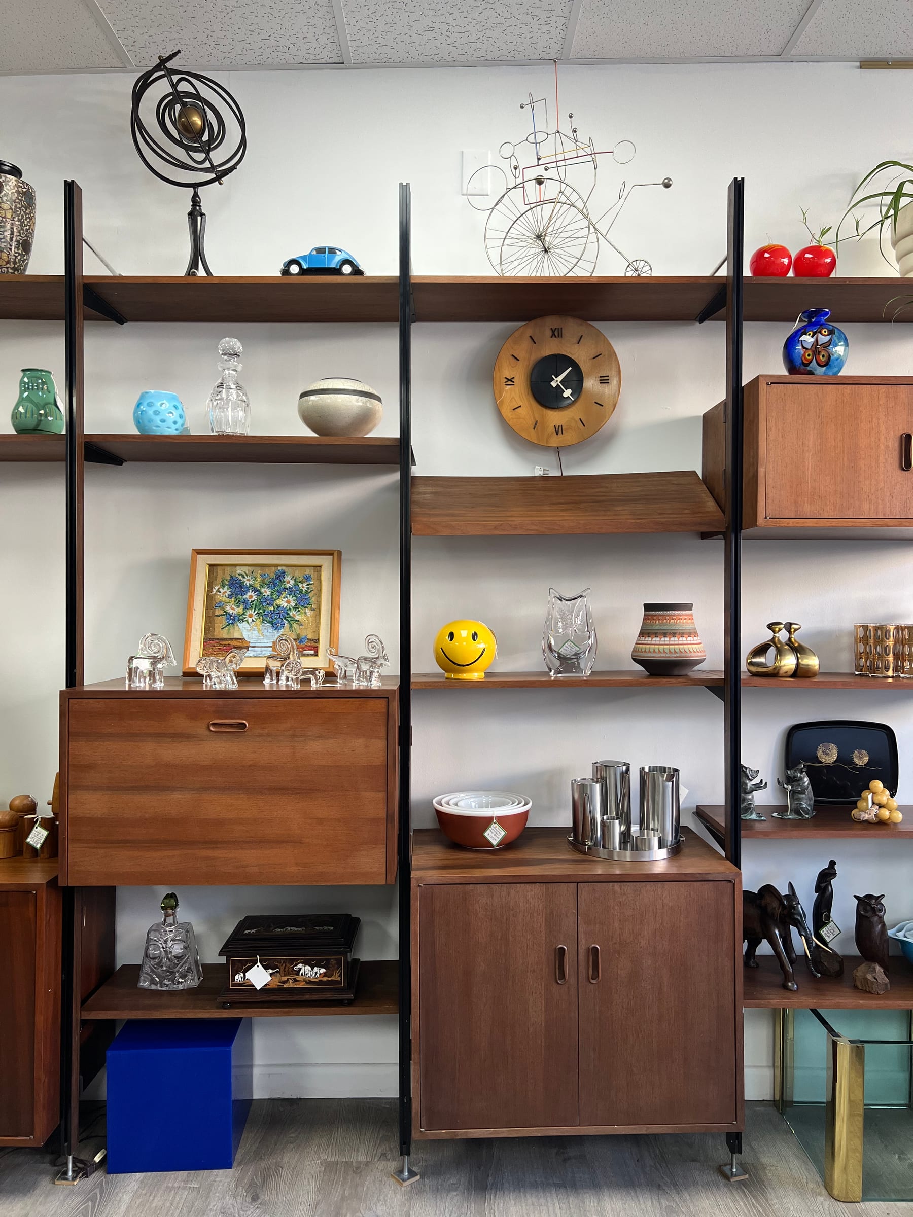 A walnut wall unit holding a vintage atomic clock, a kinetic sculpture, ceramic vases, and a midcentury landscape painting.