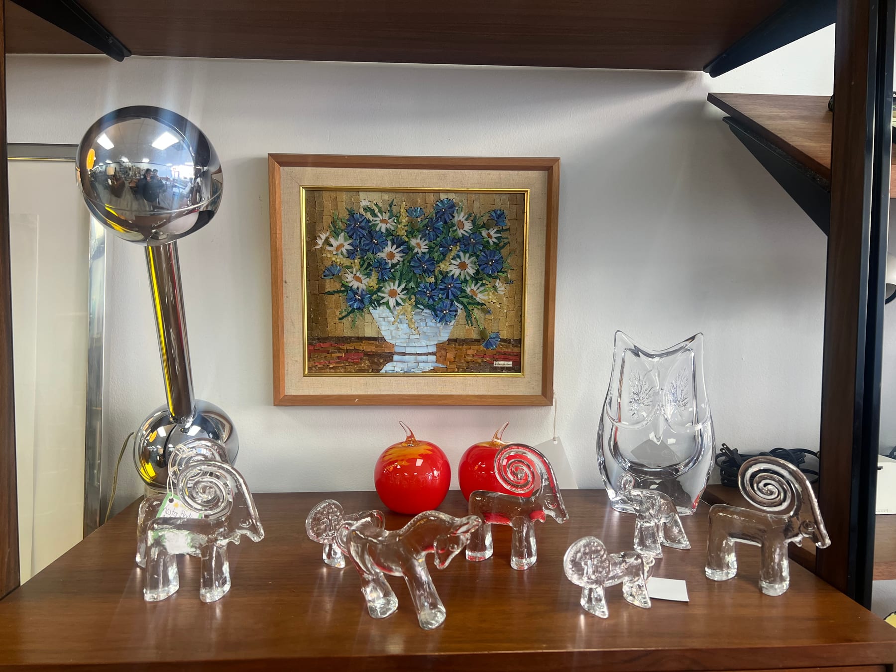 A row of clear-glass elephant figurines in front of a chrome ball lamp and a framed floral oil.