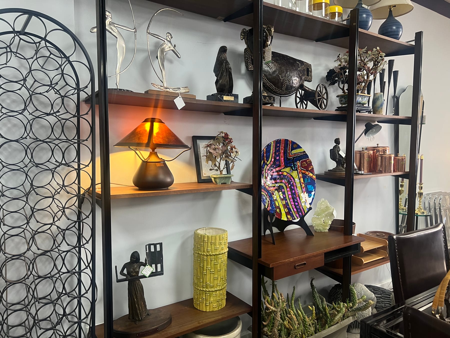 A floor-to-ceiling shelving unit with bronze sculptures, brass-and-glass lamps, framed art, and a hand-painted decorative plate.