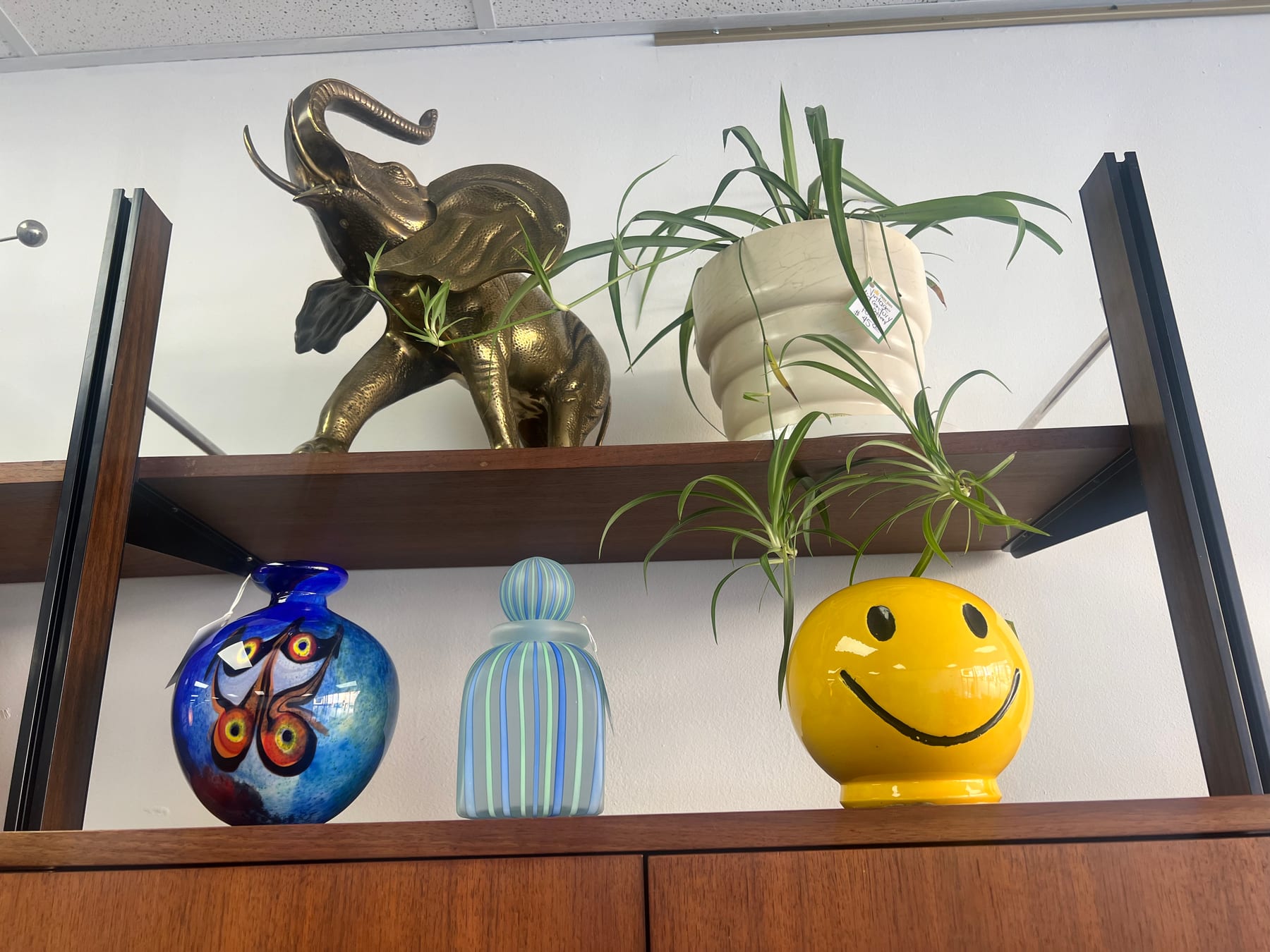 A brass elephant figurine on the upper shelf next to a yellow smiley-face planter and a hand-painted blue glass urn.